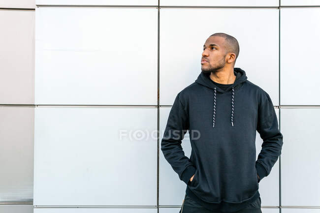 Cool African American male in casual wear with hands in pockets looking up in city — Fotografia de Stock
