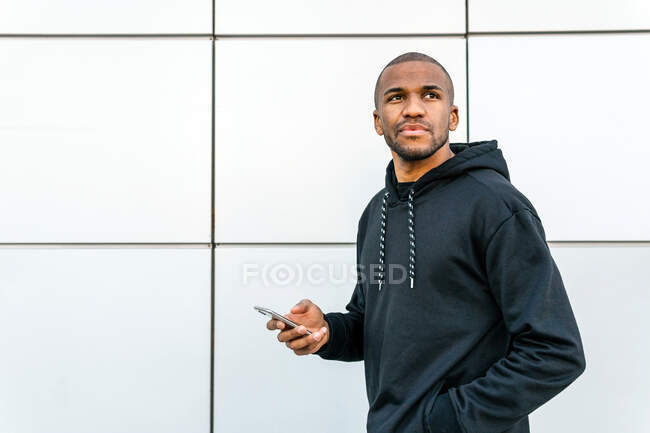 Cool hombre afroamericano en ropa casual de navegación en el teléfono móvil mientras mira hacia otro lado de pie en cerca de la pared de azulejos en la ciudad - foto de stock