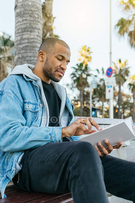 Вид со стороны взрослого бородатого этнического мужчины в повседневной одежде, читающего журнал, сидя на городской скамейке — стоковое фото