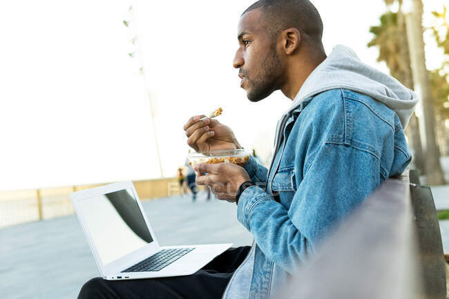 Side view of crop dreamy ethnic male distance worker with delicious lunch in container sitting with netbook in city and looking away — Stock Photo
