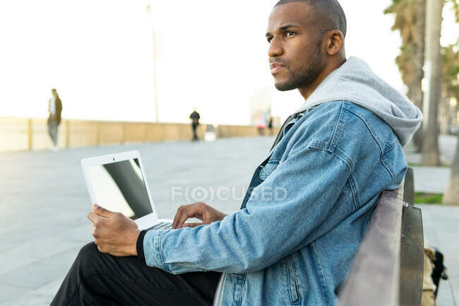 Side view of ethnic African american adult male remote employee with laptop sitting in city — Stock Photo