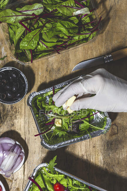 Top view crop anonymous chef in glove adding butter cubes to fresh mix leaves salad in foil bowl placed on table near cut onion and olives — Photo de stock