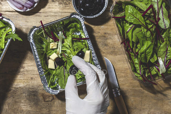 Top view crop anonymous chef in glove adding black olives to mix leaves salad with butter cubes — Stock Photo