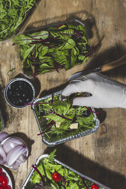Top view crop anonymous chef in glove adding black olives and leaves to mix leaves salad with butter cubes — Stock Photo