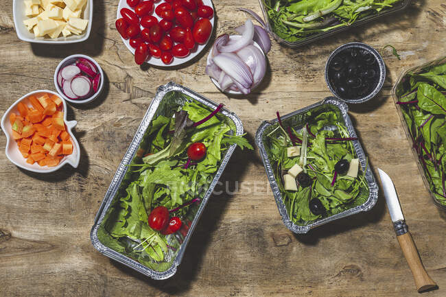 Top view composición de deliciosas ensaladas de verduras en cuencos de papel de aluminio colocados en la mesa cerca de varios ingredientes, incluyendo tomates cereza cebolla rábano y zanahorias - foto de stock