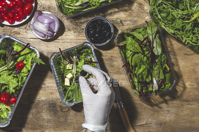 Top view chef anónimo en guante añadiendo aceitunas negras para mezclar ensalada de hojas con cubitos de mantequilla - foto de stock
