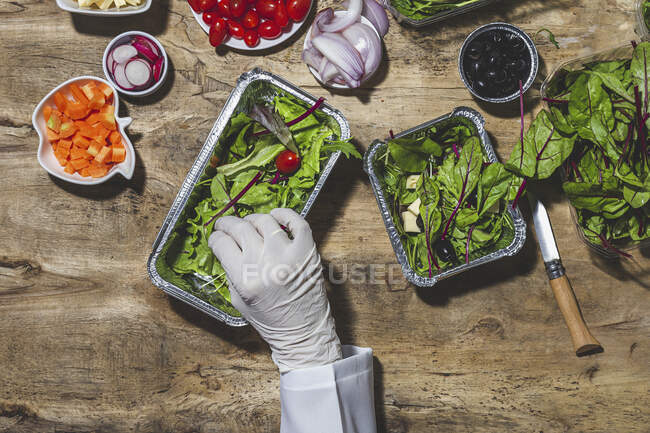 From above crop anonymous professional chef in glove adding ripe red cherry tomatoes to fresh mixed leaves in foil bowl placed on table near salad vegetable ingredients — Photo de stock