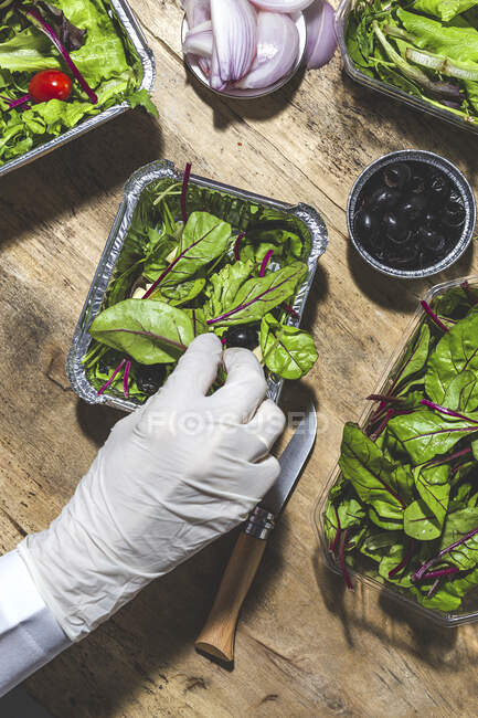 Top view coltivare anonimo chef in guanto l'aggiunta di olive nere per mescolare insalata foglie con cubetti di burro — Foto stock