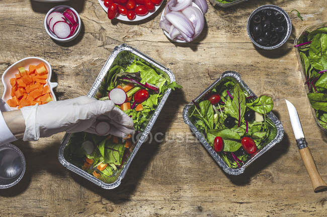 From above crop anonymous professional chef in glove adding onion slices to fresh mixed leaves in foil bowl placed on table near salad vegetable ingredients — Photo de stock