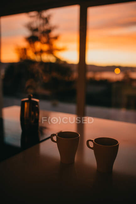 Кружки и гейзер кофеварка с горячим напитком размещены на столе в доме на фоне захватывающего заката неба — стоковое фото