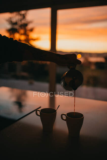 Обрізати анонімну людину, що виливає свіжий напій з гейзера кавоварки в кубки на фоні дивовижного заходу сонця — стокове фото