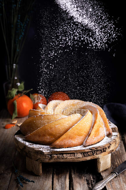 Аппетитный сладкий домашний торт посыпанный белым сахарным порошком подается на деревенском деревянном столе со свежими мандаринами — стоковое фото