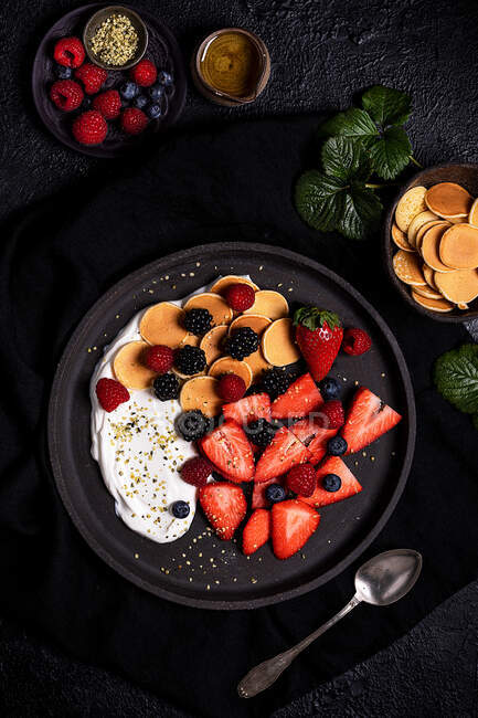 Верхний вид вкусный здоровый домашний завтрак со свежими сортами ягод подается с йогуртом и печеньем в черной тарелке на столе с ингредиентами — стоковое фото
