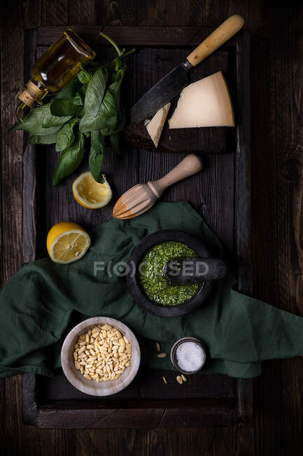 Composición de vista superior con ingredientes para la salsa tradicional de pesto incluyendo queso parmesano y hojas de albahaca y limón y piñones dispuestos en mesa oscura - foto de stock