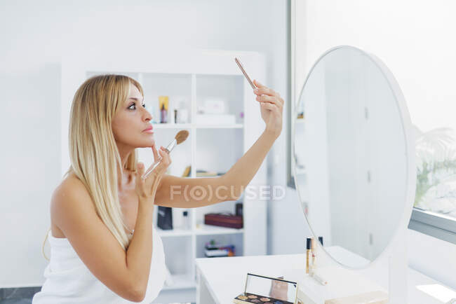 Vista lateral da fêmea com a pele perfeita aplicando pó e rosto tocando ao tirar auto-tiro no smartphone enquanto faz maquiagem em casa — Fotografia de Stock