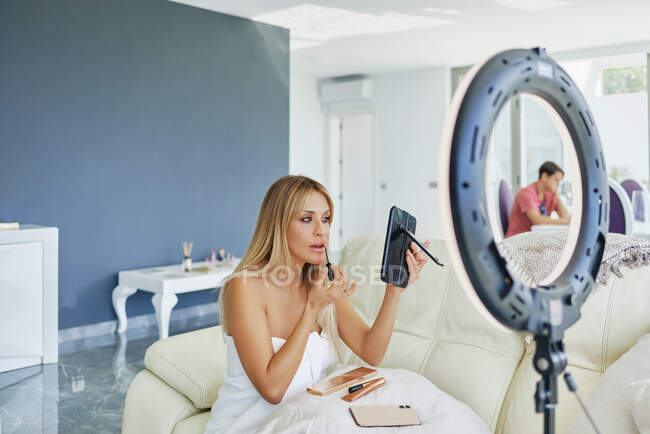 Vista lateral de la hembra aplicando brillo de labios y mirándose en el espejo mientras está sentado en la habitación con lámpara de círculo - foto de stock