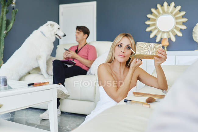 Mulher pacífica aplicando lápis de sobrancelha enquanto faz maquiagem no quarto com cachorro fofo e namorado — Fotografia de Stock