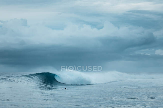Persona irreconocible nadando en la tabla de surf en poderosas olas marinas espumosas rodando y salpicando sobre la superficie del agua contra el cielo azul nublado - foto de stock