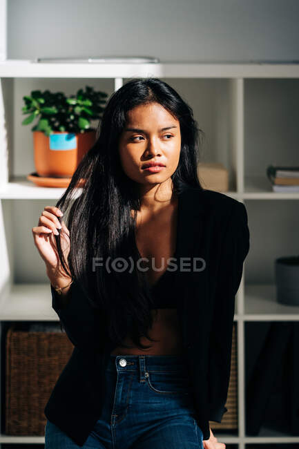 Молодая привлекательная латиноамериканка в черной куртке и джинсах сидит на стуле в современной светлой комнате и смотрит в сторону — стоковое фото