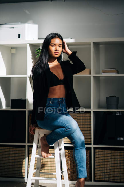 Young attractive Hispanic woman wearing black jacket and jeans sitting on chair in modern light room and looking at camera — Fotografia de Stock