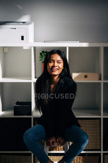 Fröhliche junge hispanische Frau in schwarzer Jacke und Jeans sitzt auf Stuhl in modernem hellen Raum und schaut weg — Stockfoto