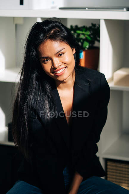 Cheerful young Hispanic woman wearing black jacket and jeans sitting on chair in modern light room and looking at camera — Photo de stock