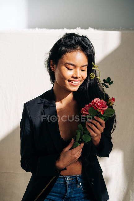 Young charming ethnic female model wearing black robe touching hair while looking at pink roses bouquet in shadow from sunlight — Photo de stock