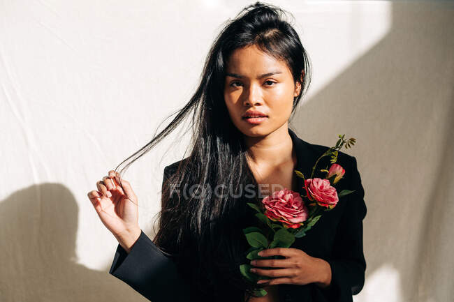 Junges bezauberndes ethnisches Model in schwarzer Robe, das die Haare berührt, während es in die Kamera blickt und rosa Rosen im Schatten des Sonnenlichts hält — Stockfoto