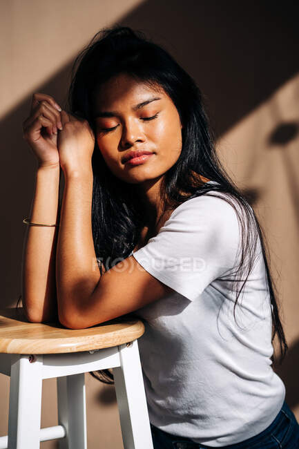 Full body of confident young Hispanic lady wearing casual clothes standing on knees and leaning on wooden chair in shadow from sunlight — Fotografia de Stock