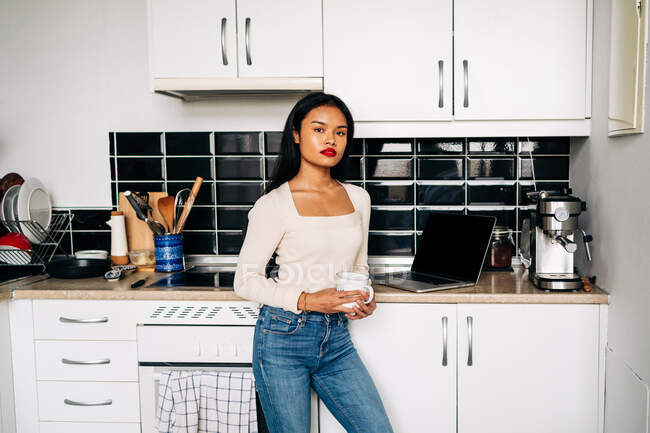Confident young Hispanic lady wearing casual outfit standing near kitchen counter with portable laptop and looking at camera while carrying ceramic cup — Stock Photo
