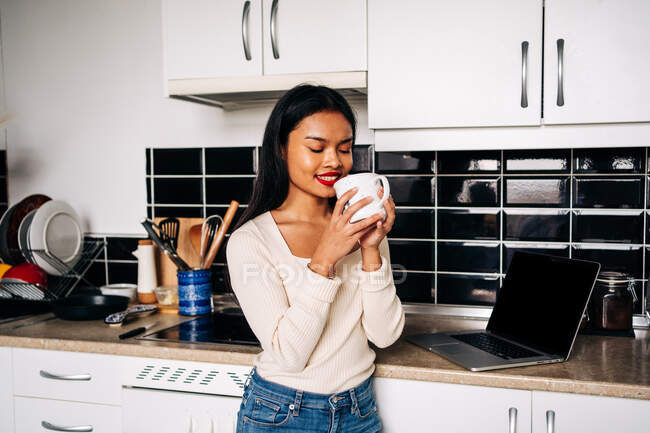 Giovane donna ispanica sorridente che indossa abbigliamento casual in piedi vicino al bancone della cucina con laptop portatile con gli occhi chiusi in possesso di tazza di ceramica — Foto stock