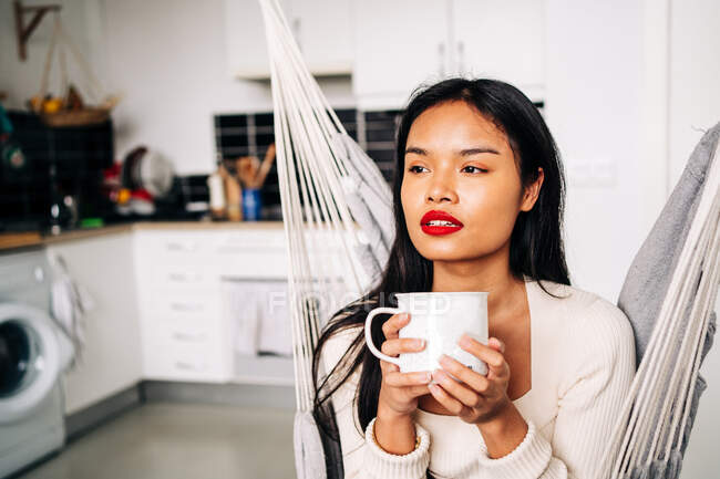 Pensive young Hispanic woman sitting in hammock in modern kitchen with hot beverage during daytime — Photo de stock