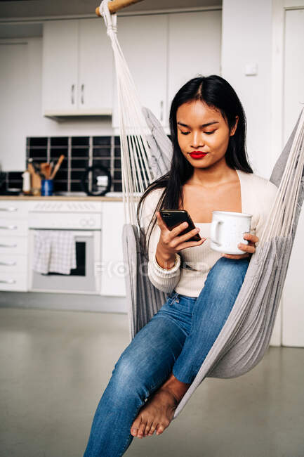 Mujer hispana joven infeliz sentada en hamaca en cocina moderna con bebida caliente y usando un teléfono inteligente moderno durante el día - foto de stock