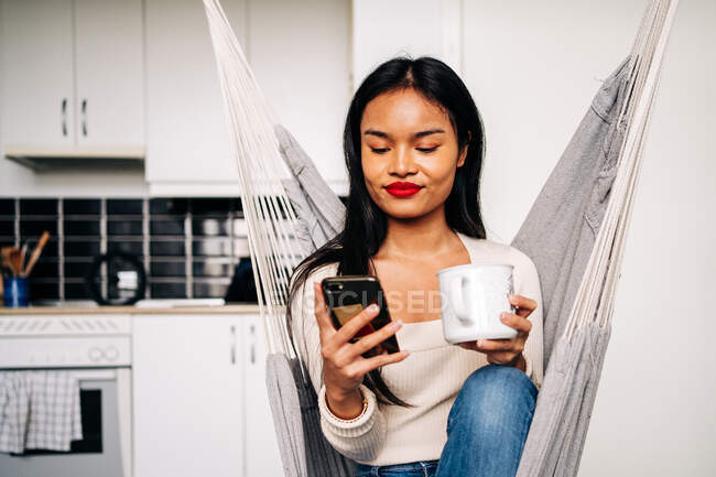 Young Hispanic woman sitting in hammock in modern kitchen with hot beverage and using modern smartphone in daytime — Photo de stock