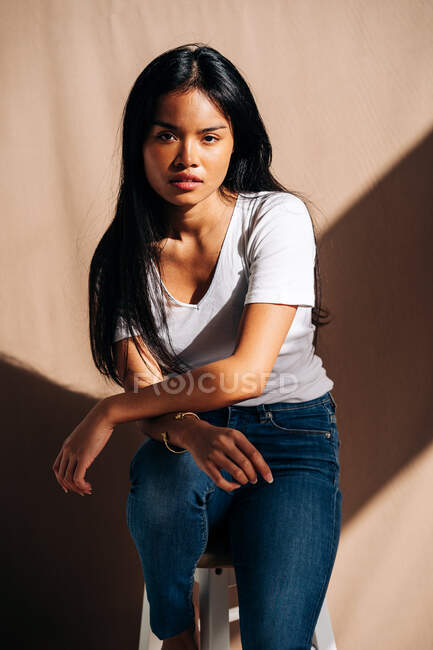 Full body of confident young Hispanic lady wearing casual clothes standing on knees and leaning on wooden chair in shadow from sunlight — Fotografia de Stock