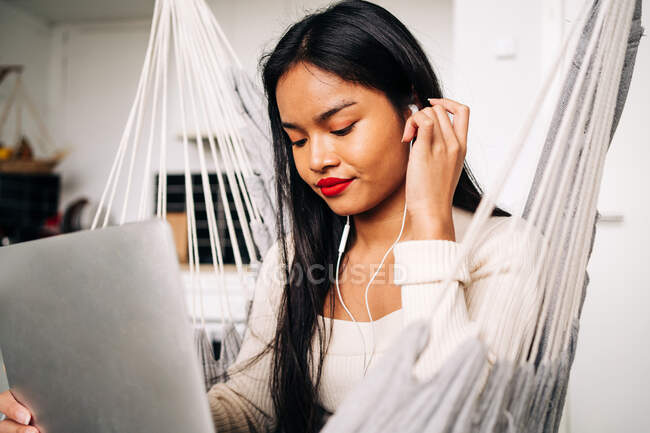 Mujer joven sonriente con el pelo largo oscuro usando el touchpad del ordenador portátil con los auriculares mientras que se sienta en hamaca adentro - foto de stock