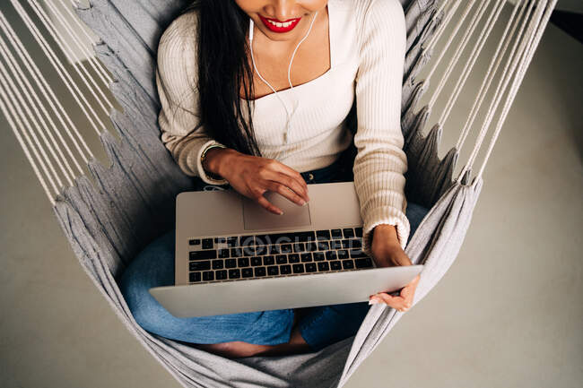 From above of crop smiling young woman with long dark hair using touchpad of portable laptop with earphones while sitting in hammock indoors — Photo de stock
