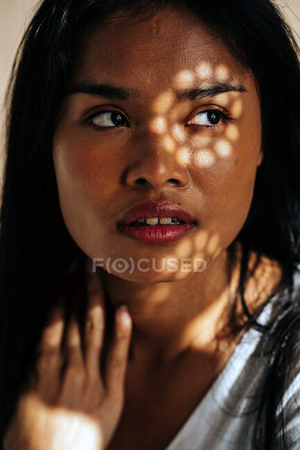 Attractive young Hispanic woman wearing white cloth touching neck while looking away in sun shadow — Fotografia de Stock