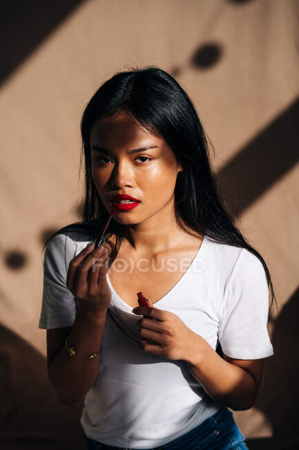 Portrait of thoughtful ethnic female with long dark hair looking at camera and rouging lips with red lipstick — Stock Photo