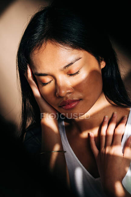 Attractive young Hispanic woman wearing white cloth touching neck with eyes closed in sun shadow — Stock Photo