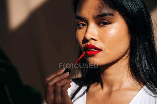 Portrait of crop thoughtful ethnic female with long dark hair looking at camera and rouging lips with red lipstick — Photo de stock