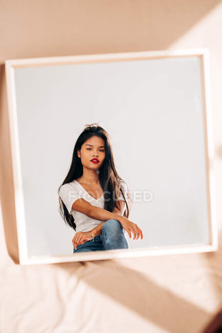 Reflection of pensive ethnic female in casual outfit with long hair and bright lips looking in mirror — Photo de stock
