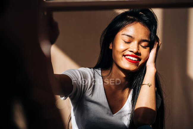 Cheerful ethnic female in white t shirt with red lips with eyes closed and stretching out hand — Photo de stock