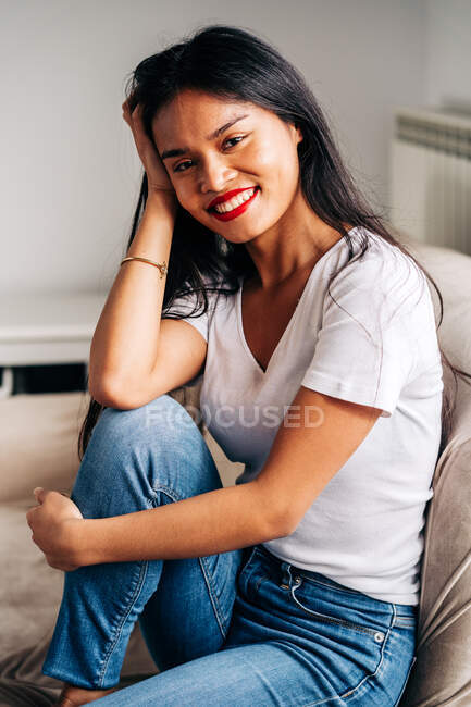 Портрет на веселой этнической женщине, сидящей на мягком диване и смотрящей в камеру в свободное время — стоковое фото