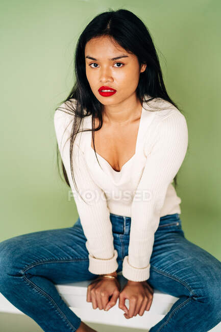 Привлекательная модель этнической женщины с яркими губами, смотрящей в камеру и опирающейся на руки, сидящей в студии на зеленом фоне — стоковое фото