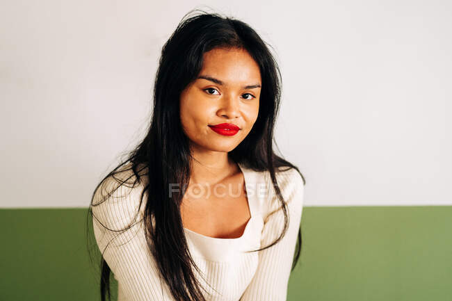 Attraente modello etnico femminile con labbra luminose guardando la fotocamera seduta in studio su sfondo verde — Foto stock