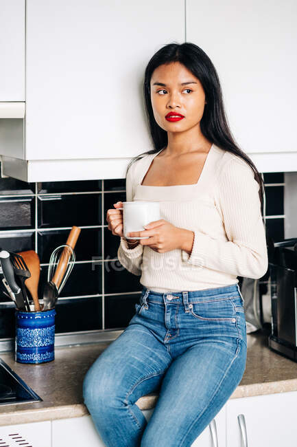 Wistful ethnic female in casual clothes sitting on countertop with cup of coffee while resting at home — Fotografia de Stock
