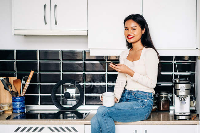 Усміхнена етнічна жінка в повсякденному одязі сидить на стільниці з чашкою перегляду кави на мобільному телефоні, відпочиваючи вдома — стокове фото
