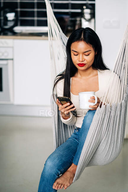 Молода іспаномовна жінка сидить у гамаку на сучасній кухні з гарячим напоєм і користується сучасним смартфоном вдень. — стокове фото