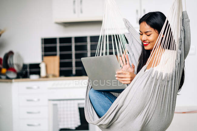 Mujer joven sonriente con el pelo largo oscuro usando el touchpad del ordenador portátil con los auriculares mientras que se sienta en la hamaca adentro que mira hacia otro lado - foto de stock